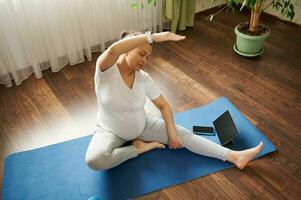 parte superior ver de un embarazada mujer haciendo yoga en un azul ejercicio estera en frente de digital artilugio, repitiendo después entrenador en línea foto