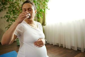 africano americano bonito embarazada mujer participación su barriga, disfrutando té ceremonia después prenatal aptitud ejercicios a hogar foto