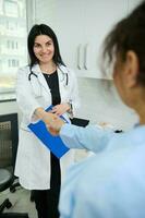 Charming pleasant female doctor holding clipboard, smiling, shaking hand and welcoming a pregnant patient in her office photo