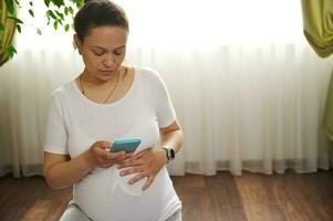 africano americano hermosa adulto mujer conmovedor su embarazada barriga, utilizando móvil teléfono durante el embarazo yoga práctica foto