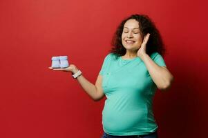 Studio portrait of curly multi ethnic beautiful happy pregnant woman holding knitted blue baby booties, isolated on red photo