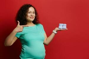 Charming pregnant woman points aside cute baby booties on her hand, smiles looking at camera, isolated on red background photo