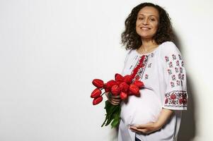 encantador adulto embarazada mujer en ucranio étnico bordado camisa, sonriente, acariciando barriga, posando con rojo tulipanes foto