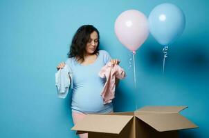 embarazada mujer, expectante madre tomando fuera azul y rosado recién nacido monos desde un cartulina caja en género revelar fiesta foto