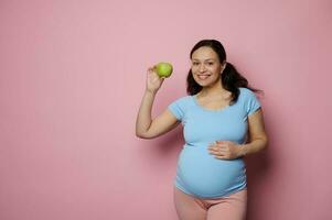 hermosa multiétnico embarazada mujer sonriente a cámara, conmovedor su barriga, participación un manzana, aislado rosado antecedentes foto