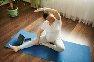 embarazada mujer practicando en línea el embarazo yoga en un estera, haciendo prenatal extensión en cuerpo equilibrar ejercicio. parte superior ver foto