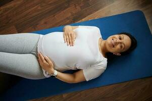 Top view pregnant woman caresses her belly, smiles at camera during prenatal relaxation exercises on fitness mat at home photo