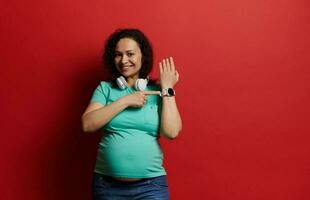 Smiling pretty woman, pregnant expectant mother looking at camera and showing her smart wrist watch, red background photo