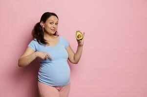 Smiling vegan expectant woman, holds one half of fresh avocado fruit, points at her pregnant belly. Healthy nutrition photo