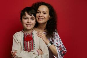 Portrait of happy mother and son wearing Ukrainian ethnic embroidered shirts, looking at camera, isolated red background photo