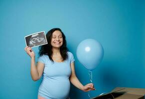 emocionado embarazada mujer expresando positivo emociones esperando bebé chico, participación bebé ultrasonografía y azul globo foto