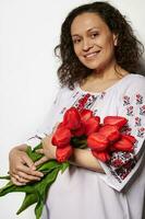 Beautiful woman in embroidered white dress, holds a bouquet of red tulips, smiles looking at camera, isolated on white photo