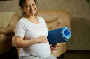 Happy pregnant woman smiles looking at camera, holds a fitness mat and gently caresses her belly, enjoying her pregnancy photo