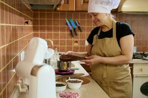 Charming housewife holding a baked chocolate biscuit soaked in strong coffee, prepares a delicious chocolate cake photo