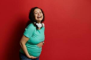 Cheerful multi ethnic pregnant woman, expectant mom holding her belly, smiling and laughing over red isolated background photo