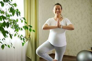 Authentic conscious pregnant woman with eyes closed, practices pregnancy yoga in tree posture with namaste hands gesture photo