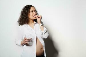 Multi-ethnic adult pregnant woman holding glass of water and pill. Healthy pregnancy, gynecology. Mother expecting baby photo