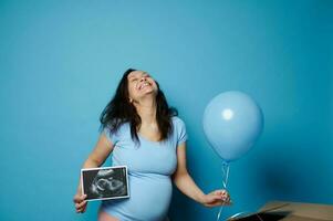 lleno de alegría embarazada mujer expresando positivo emociones esperando bebé chica, posando con azul globo y ultrasonido escanear foto