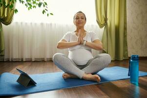 Gravid woman sitting barefoot on a fitness mat, hands palms together, deeply breathing, practicing online pregnancy yoga photo