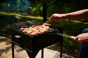 Close-up male hand pierces the marinated steak with a fork while cooking the meat on the barbecue grill in the backyard. photo