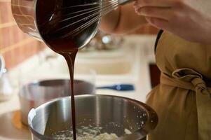 Cropped view pastry chef using whisk, adding melted confectionery dark chocolate into a bowl of food processor photo