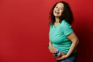 Beautiful pregnant woman laughing looking at camera, holding baby booties over her belly, isolated on red background photo