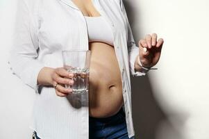 Close-up pregnant woman holding glass of water and medical pill with vitamins and minerals to take during pregnancy photo