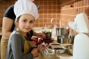 Lovely little girl in chef's hat and kitchen apron, smiling, looking at camera, helping her mother to prepare yummy cake photo