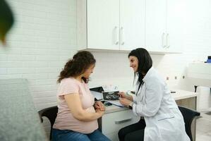 Female doctor GP, obstetrician gynecologist shows to pregnant woman ultrasound scan of her baby in medical consultation photo