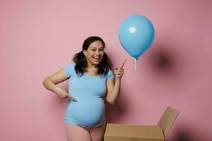 Expectant pregnant woman smiles points at blue balloon, she's expecting a baby boy, pink background. Gender reveal party photo