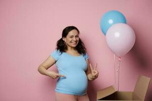 Amazed pregnant woman, expecting twins, shows two fingers looking at camera, isolated pink backdrop. Gender reveal party photo