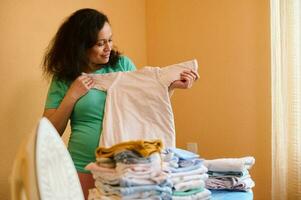 Happy pregnant woman enjoying household chores, ironing baby clothes, prepares bag for maternity hospital and childbirth photo