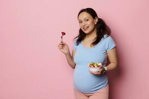 hermosa joven embarazada mujer madre, participación un cuenco con Cortado orgánico frutas, sonriente a cámara en rosado antecedentes. foto