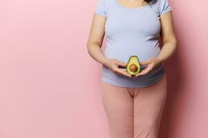 de cerca embarazada grávido mujer participación un medio de aguacate Fruta a el nivel de su grande barriga, aislado rosado antecedentes foto