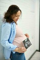 confidente étnico embarazada mujer participación el ultrasonografía de su bebé, en pie en contra blanco ladrillo pared antecedentes foto
