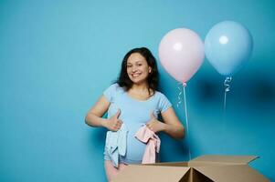 Smiling happy pregnant woman holds pink and blue baby bodysuits, thumbs up, experiencing positive emotion awaiting twins photo
