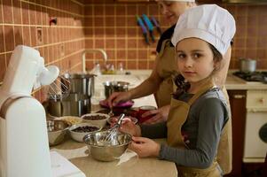 encantador niño niña en del chef sombrero y delantal, ayuda su mamá a hacer un festivo pastel mezcla azotado crema con Derretido chocolate foto