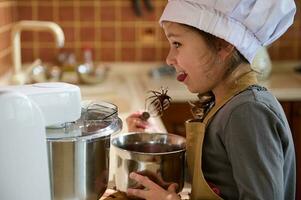 linda pequeño niña ayuda su mamá en cocina a preparar un pastel crema, tranquilamente lame un cuchara con Derretido delicioso chocolate foto