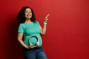 contento sonriente embarazada mujer con auriculares en su barriga, señalando dedo a Copiar espacio en rojo aislado fondo, foto