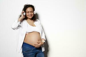 multi étnico milenario sexy grávido femenino, prenatal embarazada mujer escucha a música lista de reproducción, suavemente acariciando su barriga foto
