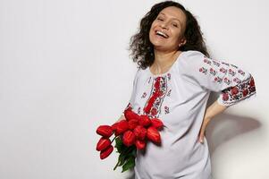 sonriente riendo contento joven embarazada mujer en bordado étnico ucranio vestido, con hermosa ramo de flores de rojo tulipanes foto