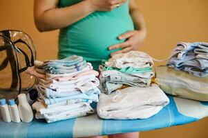 Close-up of pile of clean ironed newborn clothes and bottles with cosmetics against a pregnant woman touching her belly photo