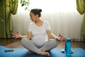 embarazada mujer acecho el embarazo yoga en línea lecciones, en móvil aplicación en su teléfono inteligente, sentado en loto actitud en estera foto