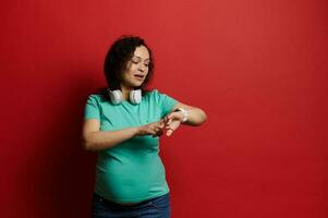 Beautiful adult pregnant woman checking her wrist watch, using new mobile app for pregnancy, isolated on red background photo