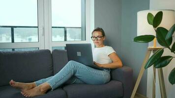 Frau mit Brille ist Sitzung auf das Couch und Arbeiten auf ein Laptop. Konzept von Fernbedienung Arbeit video