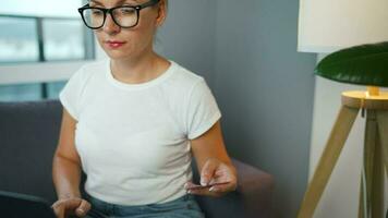 Woman with glasses sits on a couch in a cozy room and makes an online purchase using a credit card and smartphone. Online shopping video