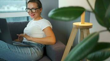 Smiling woman with glasses is sitting on the sofa and working on a laptop or chatting with someone. Concept of remote work video