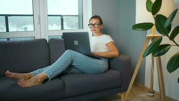 Frau mit Brille ist Sitzung auf das Couch und Arbeiten auf ein Laptop. Konzept von Fernbedienung Arbeit video