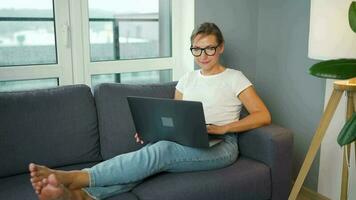 Smiling woman with glasses is sitting on the couch and working on a laptop or chatting with someone video