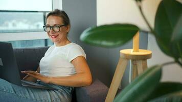 sorridente mulher com óculos é sentado em a sofá e trabalhando em uma computador portátil ou conversando com alguém. conceito do controlo remoto trabalhos video